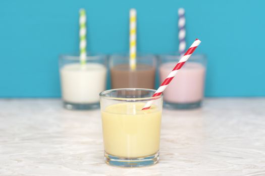 Banana milkshake with a retro paper straw in front of a row of fresh milk and flavoured milkshakes against a teal background