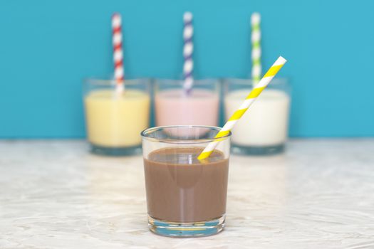 Chocolate milkshake with a retro paper straw in front of a row of flavoured milkshakes and fresh milk against a teal background