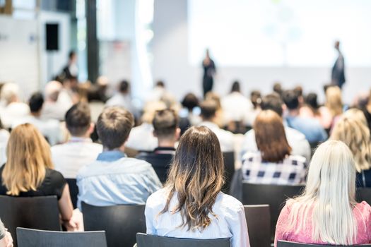 Business and entrepreneurship symposium. Speaker giving a talk at business meeting. Audience in conference hall. Rear view of unrecognized participant in audience.