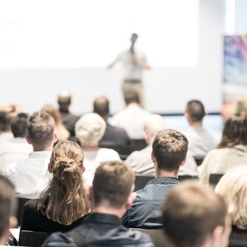 Business and entrepreneurship symposium. Speaker giving a talk at business meeting. Audience in conference hall. Rear view of unrecognized participant in audience.