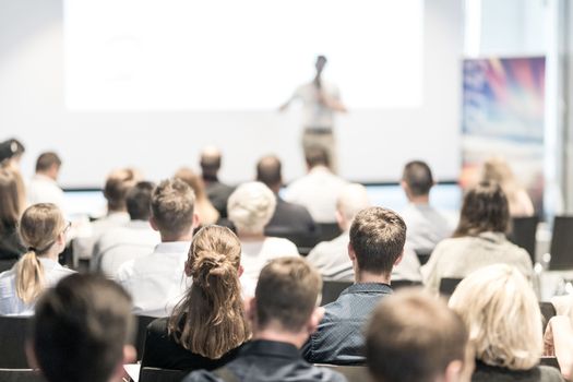 Business and entrepreneurship symposium. Speaker giving a talk at business meeting. Audience in conference hall. Rear view of unrecognized participant in audience.