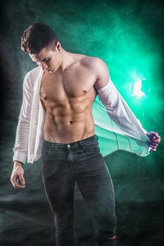 Handsome young muscular man shirtless wearing jeans, taking off leather jacket on naked muscle torso, on dark background in studio shot