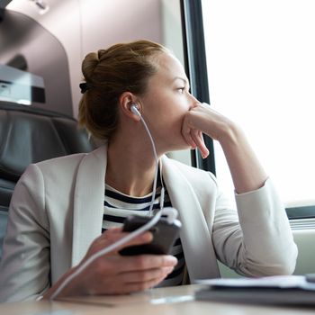Thoughtful businesswoman looking trough the window, listening to podcast on cellphone using headphone set while traveling by train in business class seat.