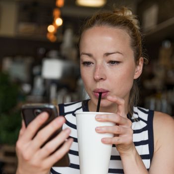 Thoughtful woman reading news on mobile phone during rest in coffee shop. Happy Caucasian female watching her photo on cell telephone while sipping coffee in cafe during free time.