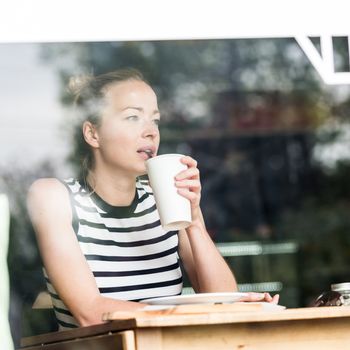 Young caucasian woman sitting alone in coffee shop drinking american coffee, people watching, thoughtfully looking trough the coffee shop window.