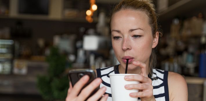 Thoughtful woman reading news on mobile phone during rest in coffee shop. Happy Caucasian female watching her photo on cell telephone while sipping coffee in cafe during free time.