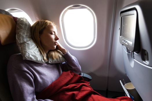 Tired blonde casual caucasian woman sleeping on seat while traveling by airplane on long distance transatlantic flight. Commercial transportation by planes.