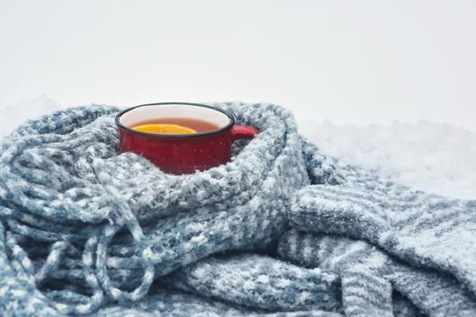 Red enameled cup of hot tea with lemon wrapped in a knitted scarf on a snowy wooden table.