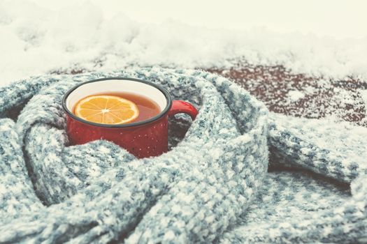 Red enameled cup of hot tea with lemon wrapped in a knitted scarf on a snowy wooden table.