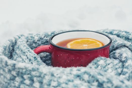 Red enameled cup of hot tea with lemon wrapped in a knitted scarf on a snowy wooden table.