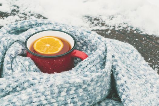 Red enameled cup of hot tea with lemon wrapped in a knitted scarf on a snowy wooden table.