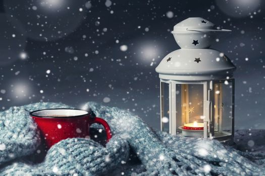 Lantern with a burning candle and a red mug with hot coffee on a snowy wooden table.