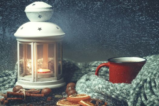 Lantern with a burning candle, spices and a red mug with hot coffee on a snowy wooden table.