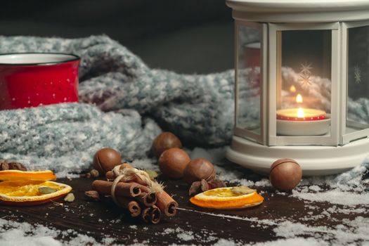 Lantern with a burning candle, spices and a red mug with hot coffee on a snowy wooden table.