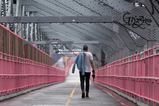 Rear view of unrecognizable stylish young man carrying jeans jacket over his shoulder walking on Williamsburg Bridge, Brooklyn, New York City, USA.
