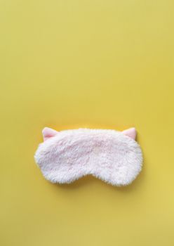Pastel pink fluffy fur sleep mask with small ears on pastel yellow paper background. Top view, flat lay, copy space. Concept of vivid dreams. Accessories for girls and young women.