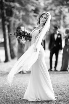 Full length portrait of beautiful sensual young blond bride in long white wedding dress and veil, holding bouquet outdoors in natural background. Black and white photo.