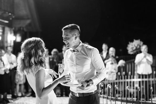 Happy bride and groom getting messy eating piece of wedding cake by their hands after cutting it. Traditional custom of wedding celebration. Black and white image.