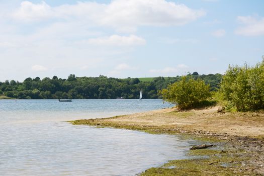 Water's edge of Bewl Water reservoir in Tunbridge Wells, Kent. An empty boat and a windsurfer are on the lake with a small ferry.