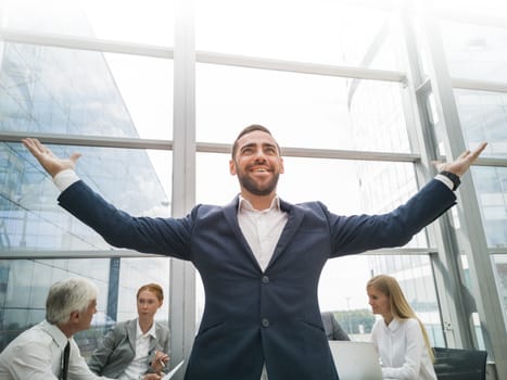 Young joyful businessman standing with arms raised smiling , team of his colleagues sitting at the table in the background