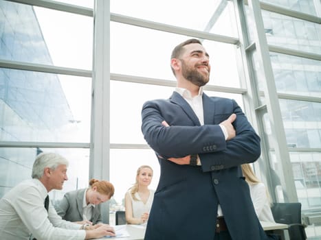 Confident young businessman standing with folded arms smiling , team of his colleagues sitting at the table in the background