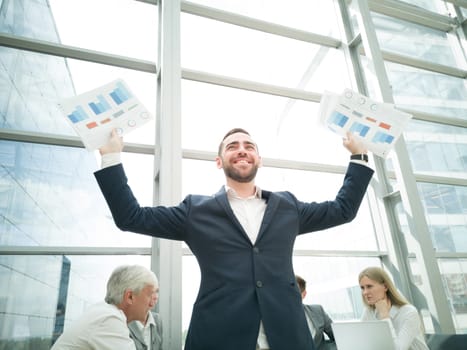Young joyful businessman standing with arms raised holding data statistics diagrams and smiling , team of his colleagues sitting at the table in the background