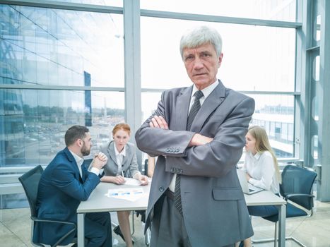 Confident mature businessman standing with folded arms smiling , team of his colleagues sitting at the table in the background