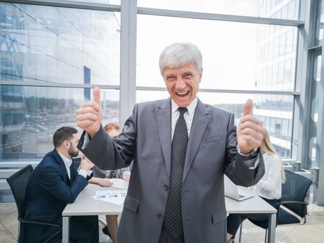 Confident mature businessman showing thumb up and smiling , team of his colleagues sitting at the table in the background