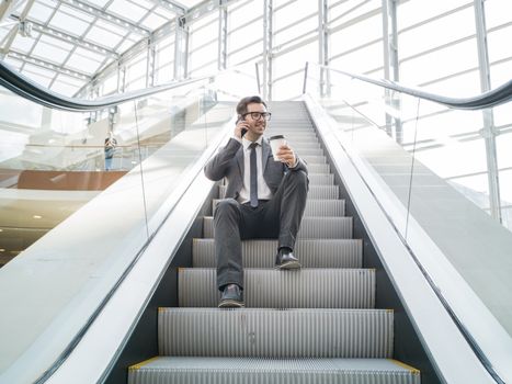 Businessman sitting on escalator talking on phone and holding coffee in hand