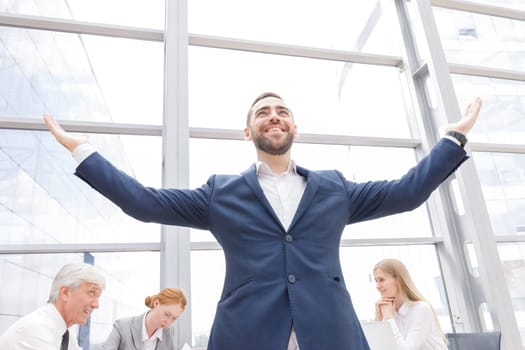 Young joyful businessman standing with arms raised smiling , team of his colleagues sitting at the table in the background