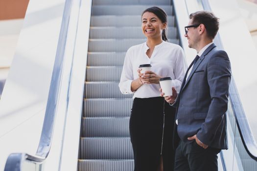 Businessman and businesswoman standing together on escalator and talk about business holding coffee in hand