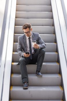 Businessman sitting on escalator surfing the net and holding coffee in hand