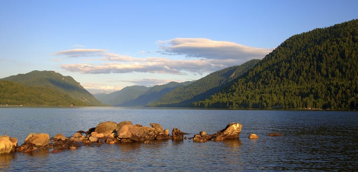 A lake surrounded by mountains at sunset and a stone ridge protruding from the shore. Teletskoye Lake, Altai, Siberia, Russia.