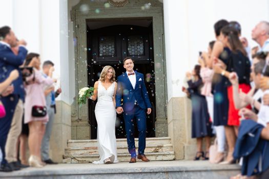 Newlyweds exiting the church after the wedding ceremony, family and friends celebrating their love with the shower of soap bubbles, custom undermining traditional rice bath.