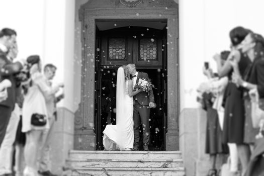 Newlyweds kissing while exiting the church after wedding ceremony, family and friends celebrating their love with the shower of soap bubbles, custom undermining traditional rice bath. Black and white.