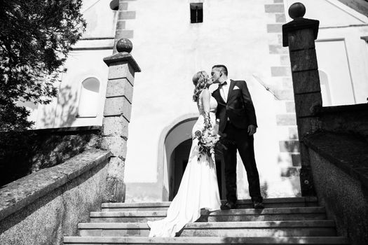 The Kiss. Bride and groom kisses tenderly on a staircase in front of a small local church. Stylish wedding couple kissing. Black and white photo.