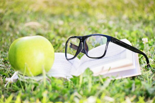 Close-up shot of glasses on a book along with an apple on a green grass. Taking a break from the studying.