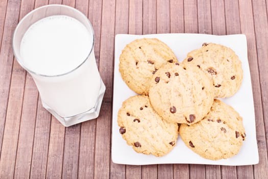 Close up shot of chocolate chip cookies on a platter with a glass of milk, on a wooden background.