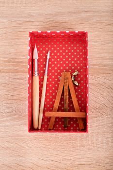 Small easel and paint brushes in a box on wooden background.