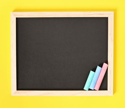 Chalkboard with wooden frame and colorful chalks on one side, isolated on yellow background.