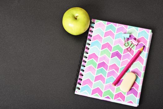 Top view of girly pink notebook with pencil, eraser, binder clips and apple on black background.