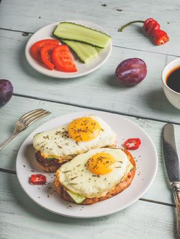 Sandwiches with vegetables and fried egg on white plate, cup of coffee and some fruits over wooden background.