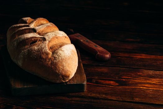 Homemade rye loaf on cutting board with knife over dark wooden background.