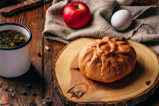 Rustic breakfast with traditional tatar pastry elesh, herbal tea in metal mug, apple and boiled eggs over dark wooden surface