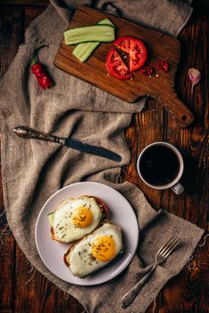 Breakfast toasts with vegetables and fried eggs on white plate and cup of coffee over grey rough cloth. View from above.