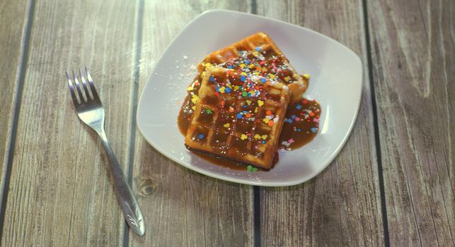 Two Viennese waffles with icing and topping lie on a plate near a fork on a wooden table.