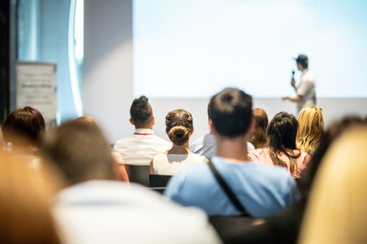 Business and entrepreneurship symposium. Speaker giving a talk at business meeting. Audience in conference hall. Rear view of unrecognized participant in audience.