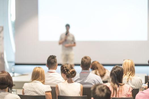 Business and entrepreneurship symposium. Speaker giving a talk at business meeting. Audience in conference hall. Rear view of unrecognized participant in audience.