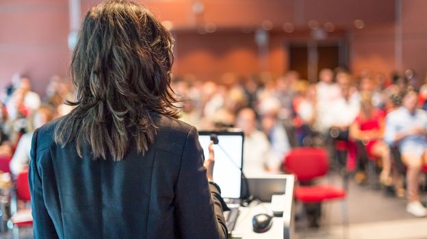 Female speaker giving a talk on corporate business conference. Unrecognizable people in audience at conference hall. Business and Entrepreneurship event.