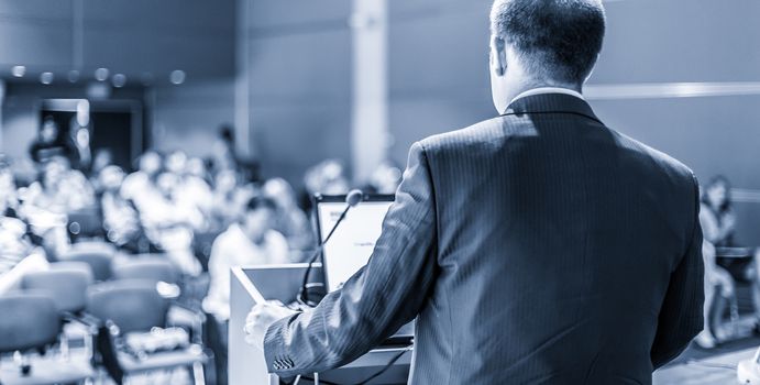 Speaker giving a talk on corporate business conference. Unrecognizable people in audience at conference hall. Business and Entrepreneurship event. Blue toned grayscale image.